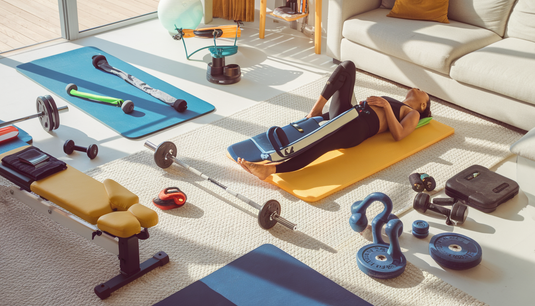 A bright home workout space with fitness equipment and a person using a compression leg massager in a sunlit living room.