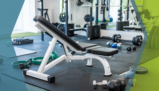 Modern home gym with a decline bench press machine surrounded by gym mats, resistance bands, barbells, and dumbbells in a vibrant blue and green color scheme.